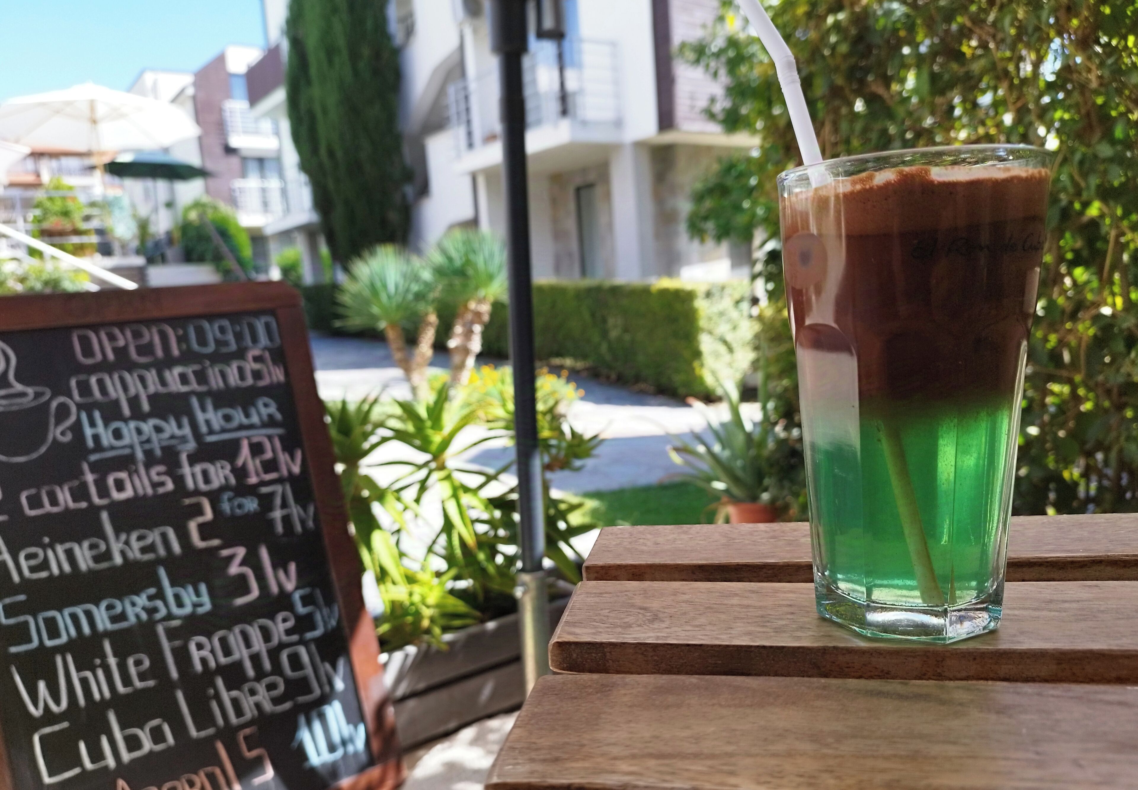 A glass of iced coffee with a straw on a wooden table, a chalkboard menu listing drinks and happy hour specials, and a building with balconies and greenery in the background.