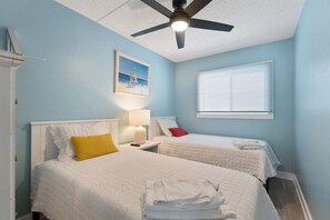 Une chambre avec deux lits, un ventilateur de plafond, une fenêtre avec des stores et un tableau au mur.