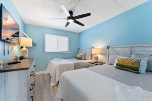 Une chambre avec deux lits, un ventilateur de plafond, des lampes de chevet, une fenêtre avec des stores et un téléviseur fixé au mur.