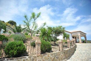 A stone-walled property with a paved entrance, a covered patio, and a variety of greenery including shrubs and trees.