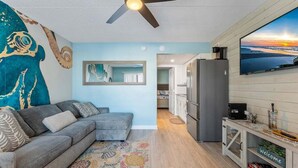 A living room with a grey sofa, a ceiling fan, a TV, and a kitchen area visible in the background. - Coastal Escape: 1 Bedroom Condo w/ 2 Heated Pools! (North Wildwood)