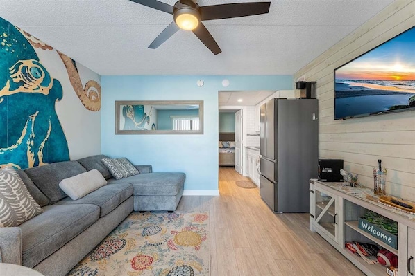 A living room with a grey sofa, a ceiling fan, a TV, and a kitchen area visible in the background.
