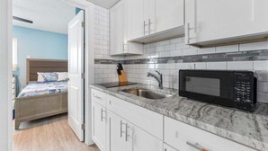 A compact kitchen with white cabinets, a sink, a microwave, and a bed in the adjacent room. - Coastal Escape: 1 Bedroom Condo w/ 2 Heated Pools! (North Wildwood)