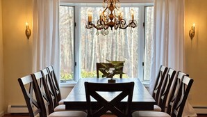 A dining room with a large wooden table, six chairs, a chandelier, and a view of trees through large windows.
