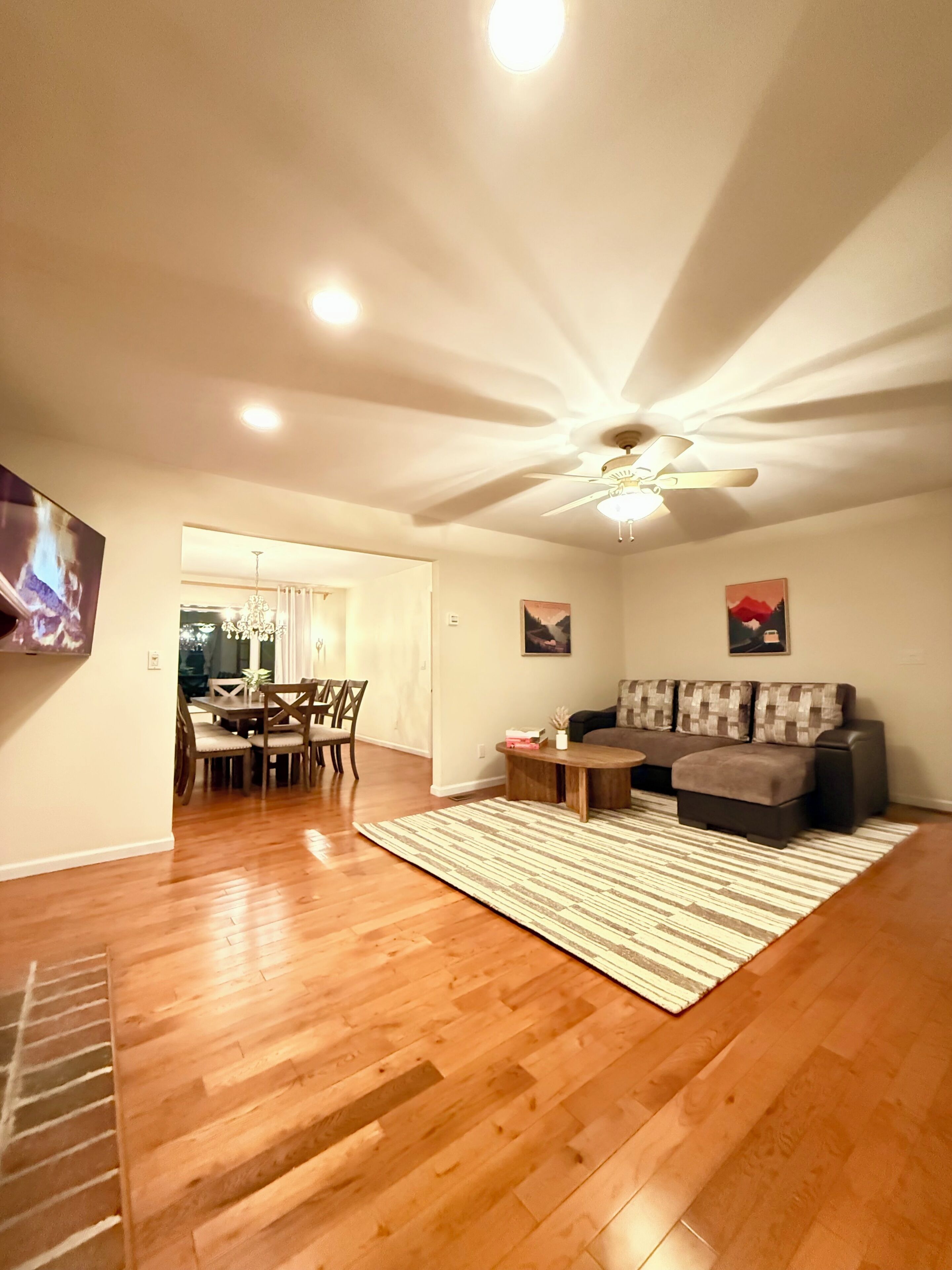 Ein geräumiges Wohnzimmer mit Holzboden, Deckenventilator, großem Flachbildfernseher, Ecksofa und Essbereich mit einer Kristallleuchtre.