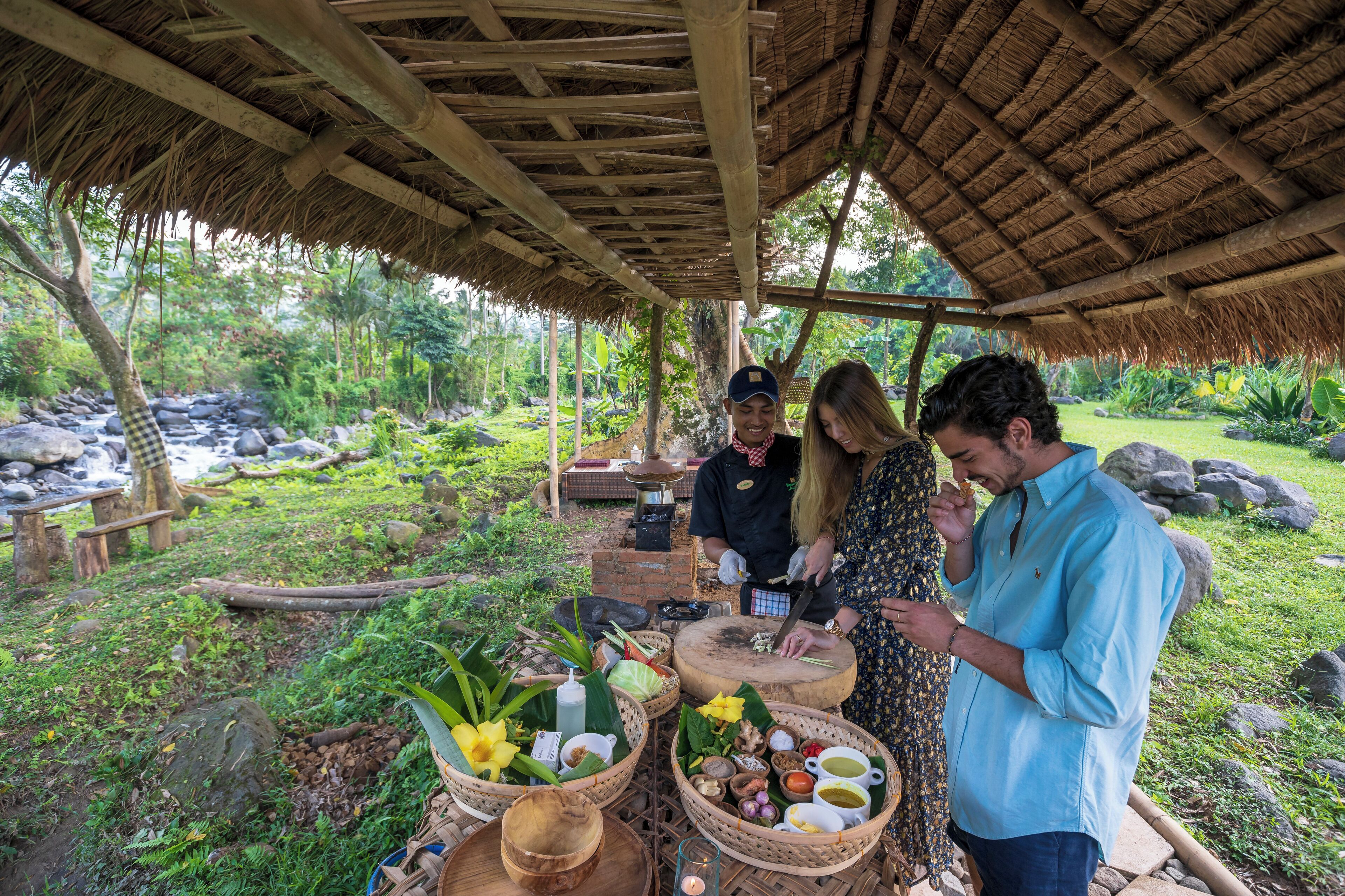 A rustic outdoor setting with a thatched roof structure, surrounded by lush greenery and a small waterfall.