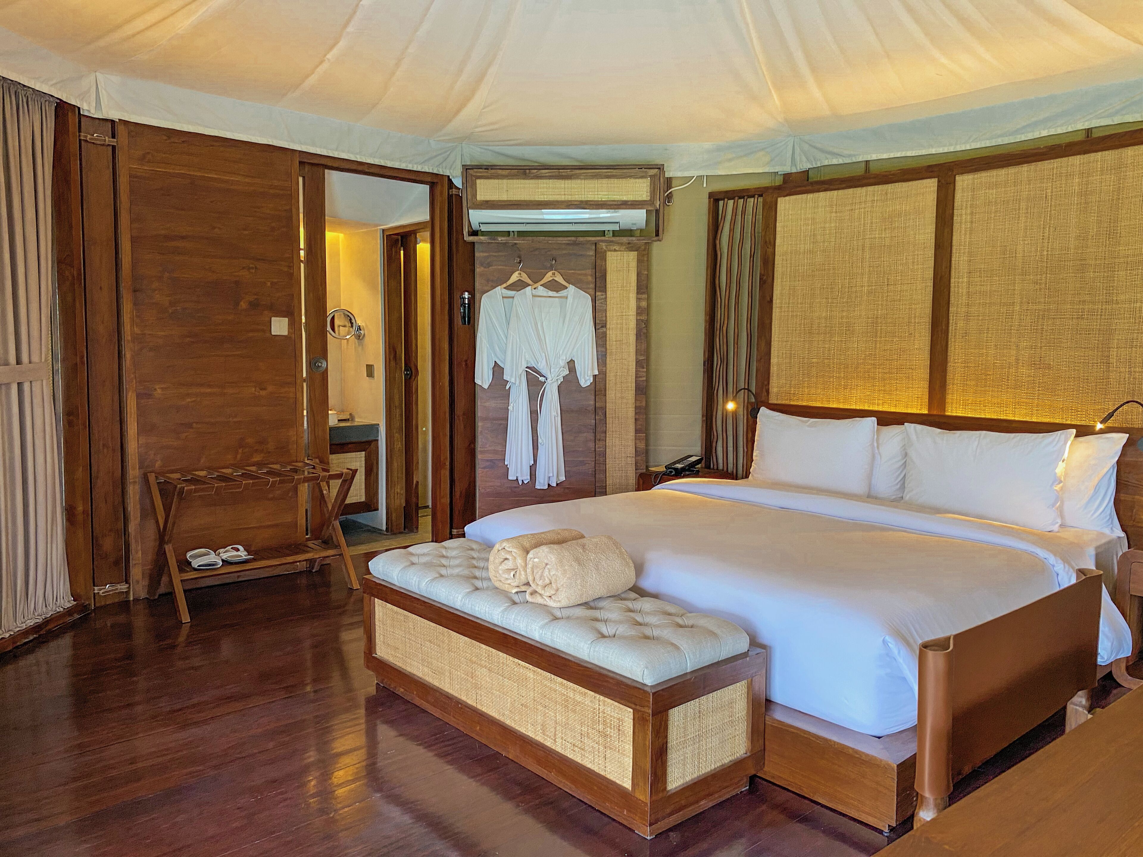 A hotel room with a bed, a wooden bench, a hanging robe, and a mirror.