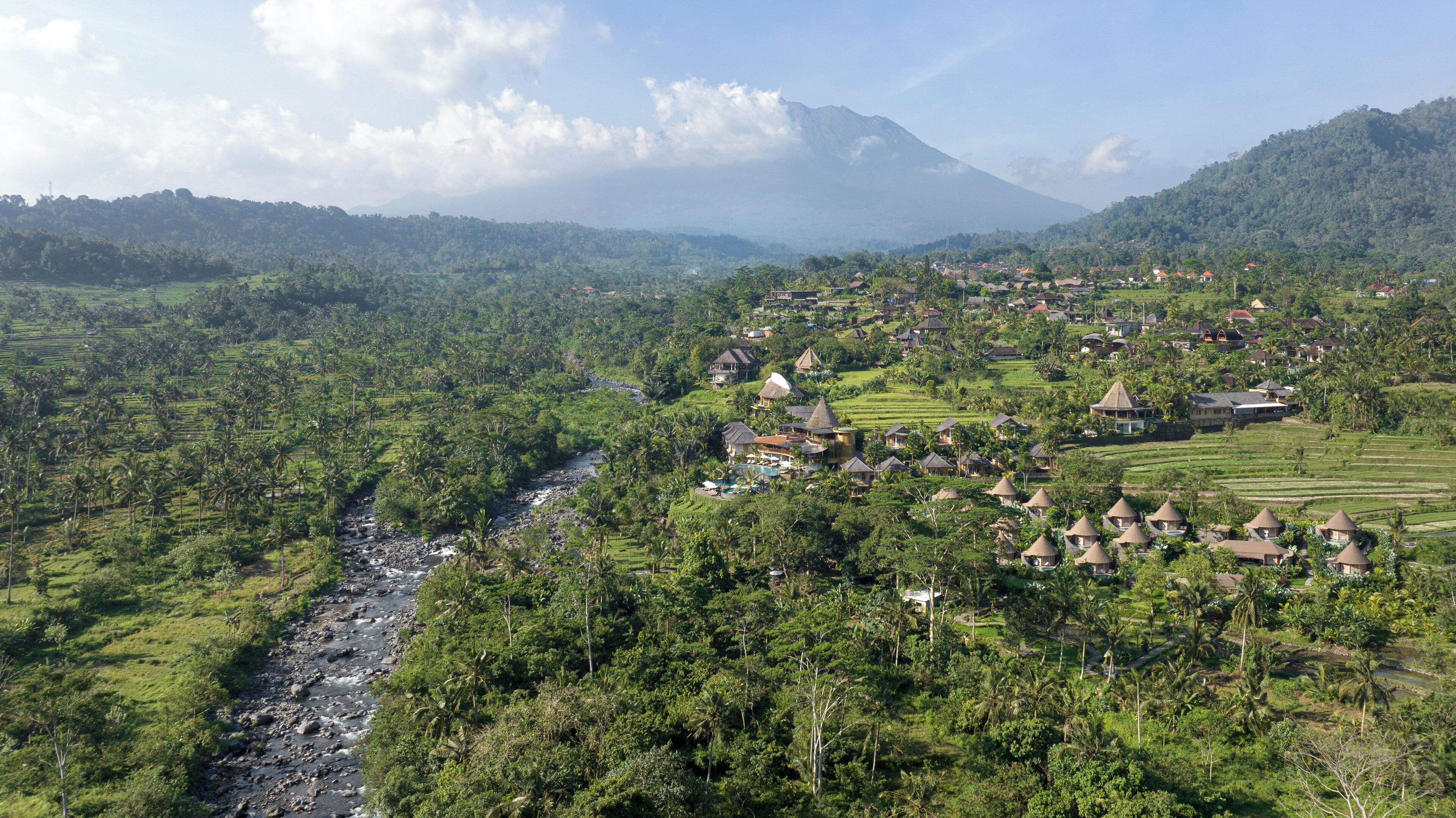A rural village with traditional thatched-roof houses, a river running through the landscape, and a mountain in the background.