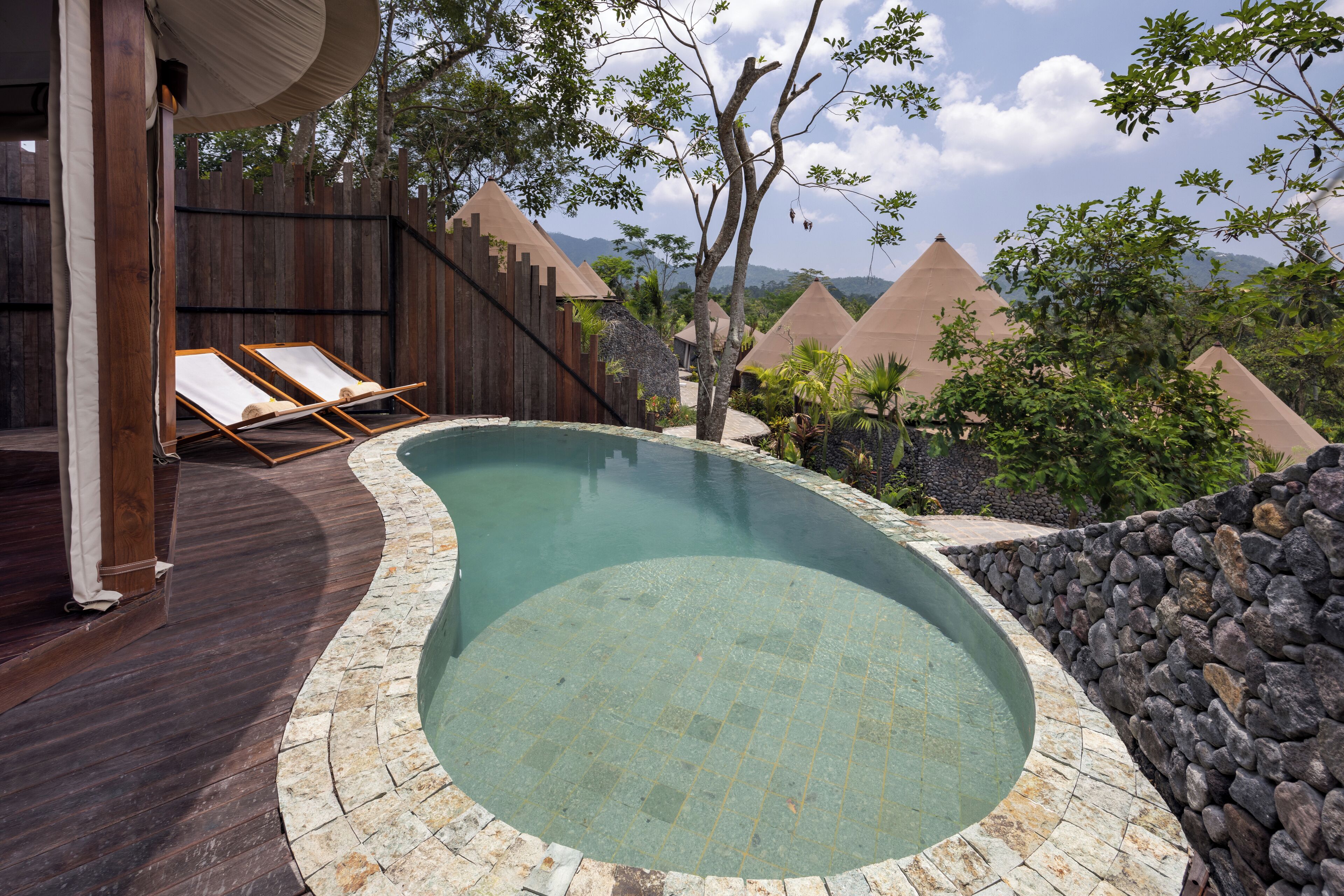 A swimming pool with a wooden deck and lounge chairs, surrounded by a stone wall and trees.