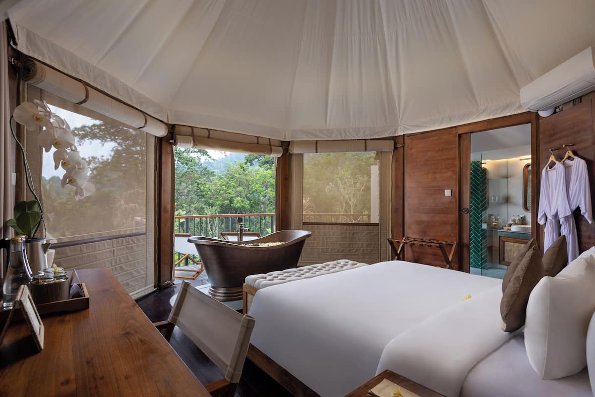Quarto com cama grande, escrivaninha de madeira, banheira e varanda com vista para o verde.