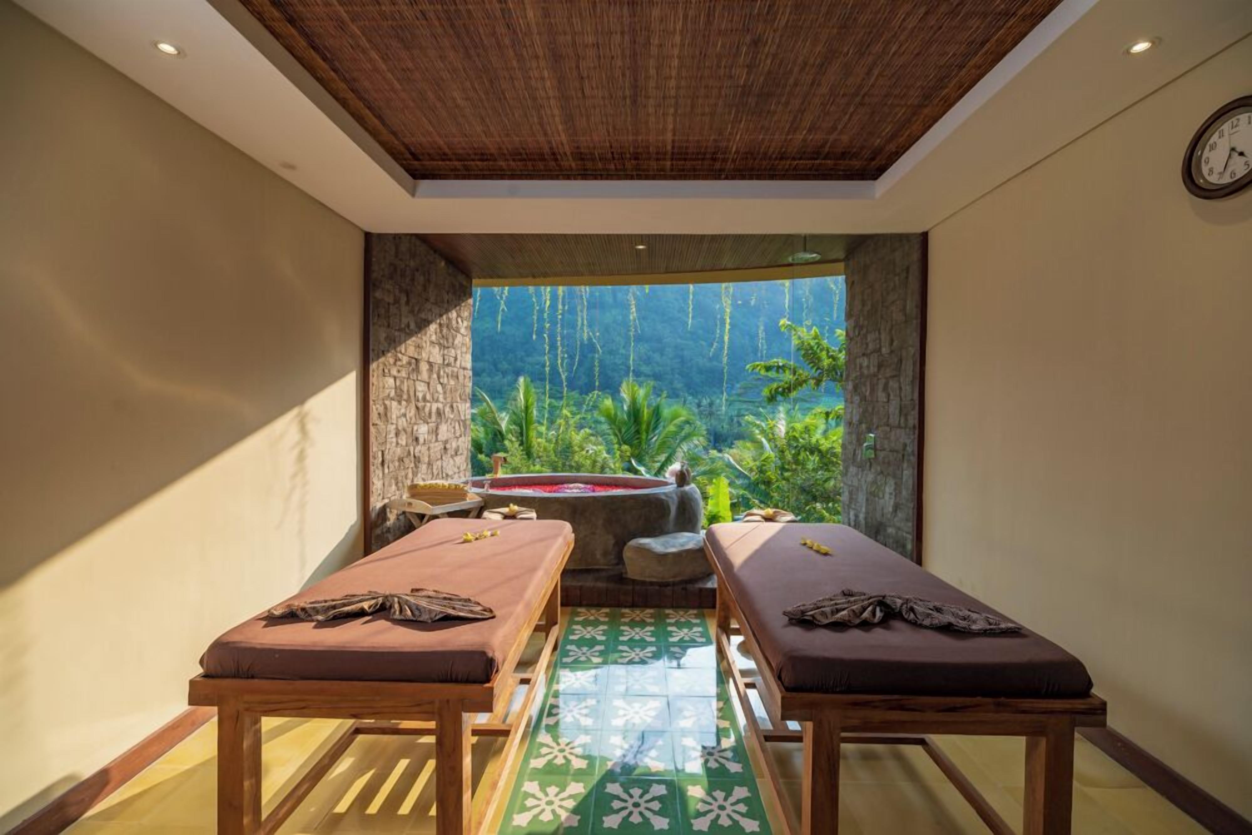 Une pièce dotée de deux tables de massage, d’un mur en pierre, d’une grande fenêtre donnant sur un paysage verdoyant et d’un plafond en bois.