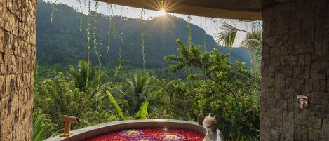 Un área de spa con un hidromasaje rodeado de flores rojas y púrpuras, una pared de piedra y vistas a vegetación exuberante y montañas.