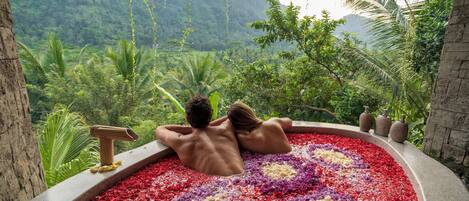 Un couple se détend dans un bain à remous, entouré d’un parterre de fleurs, avec un paysage montagneux en arrière-plan.