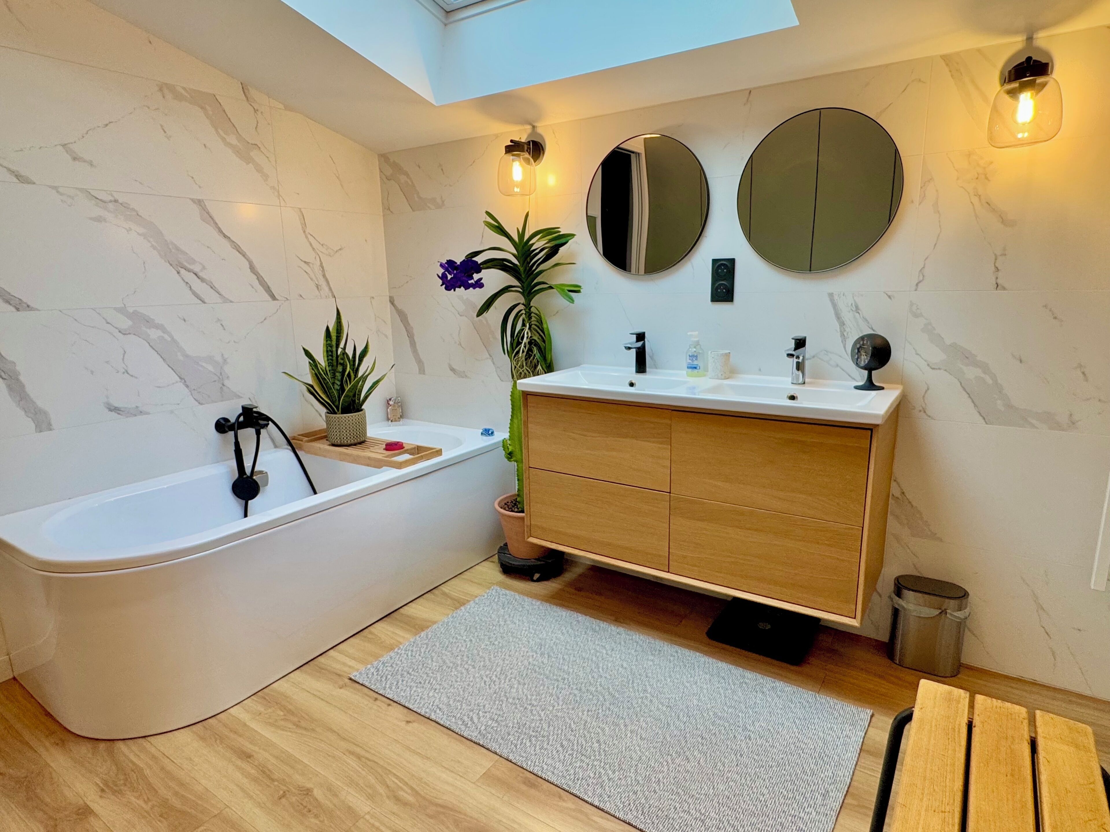 A modern bathroom with a freestanding bathtub, wooden vanity, marble walls, and a skylight.