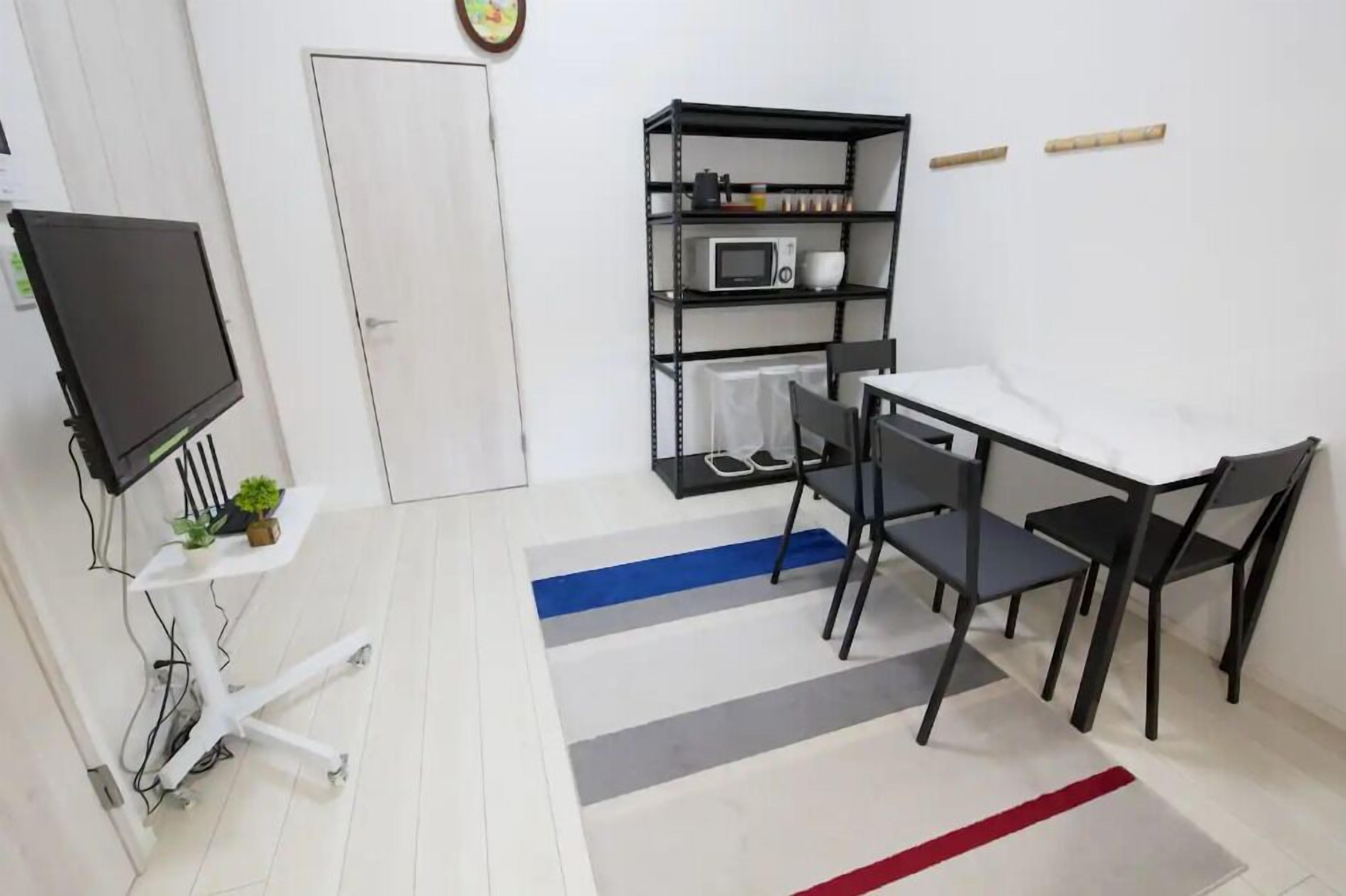 Una habitación pequeña y moderna con una puerta blanca, un televisor de pantalla plana montado en un soporte blanco, un estante metálico negro con un microondas y una mesa con cuatro sillas.