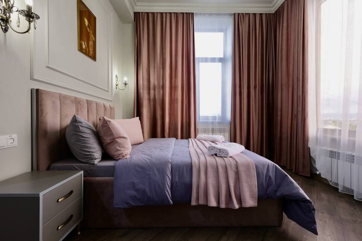 A bedroom with a bed, a nightstand, a lamp, a window with curtains, and a wall-mounted artwork.