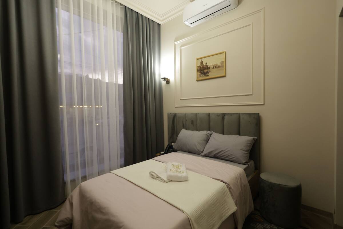 A hotel room with a bed, a bedside table, a wall-mounted air conditioner, a painting, and a window with curtains.