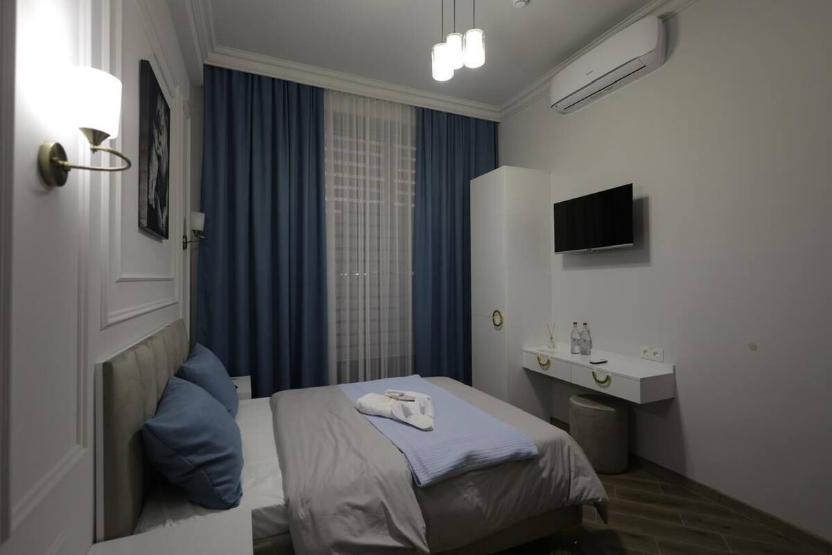 A hotel room with a bed, a bedside table with a TV, a wall-mounted air conditioner, and a window with blue curtains.