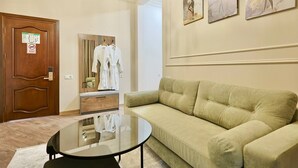 Une chambre d’hôtel avec un canapé vert, une table basse ronde en verre, une porte en bois avec une pancarte et un miroir sur lequel sont accrochés des vêtements.