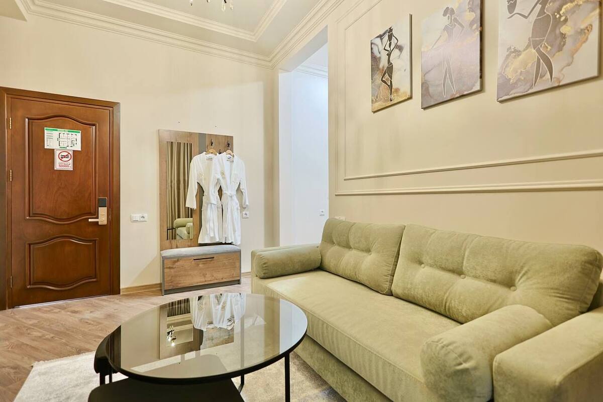 A hotel room with a green sofa, a round glass coffee table, a wooden door with a sign, and a mirror with clothes.
