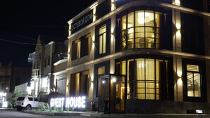 Un hôtel appelé « GUEST HOUSE », avec une enseigne devant l’entrée, de grandes fenêtres et une façade bien éclairée.