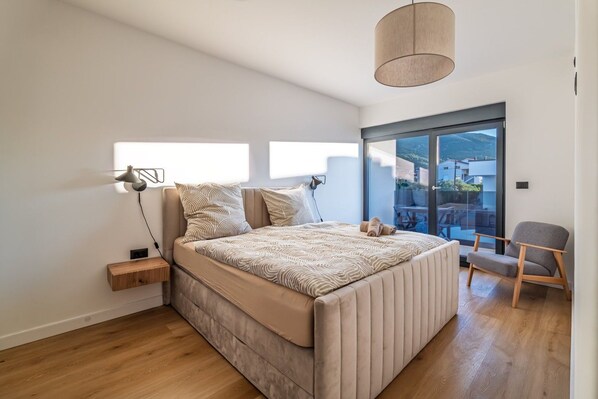 Un dormitorio moderno con una cama grande, una mesita de noche, una silla y vistas al exterior a través de un ventanal.