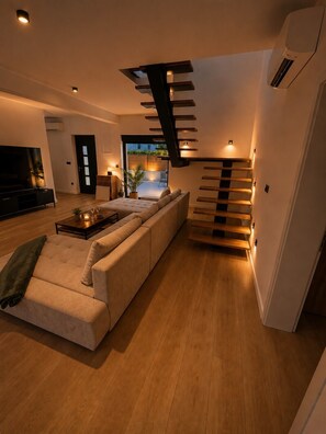 A modern living room with a sofa, a coffee table, and a TV. There is a staircase with built-in lighting.