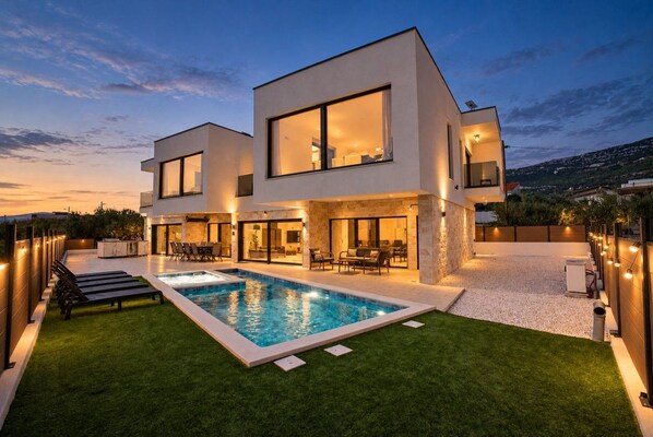 A modern two-story house with a swimming pool, outdoor seating, and a view of the surrounding landscape.