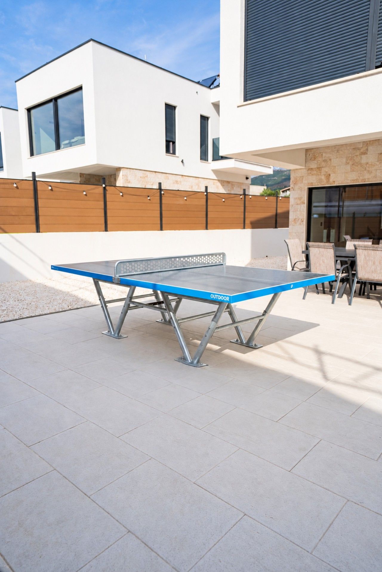 Une table de ping-pong extérieure, dotée de supports métalliques, située dans une cour avec un bâtiment moderne en arrière-plan.