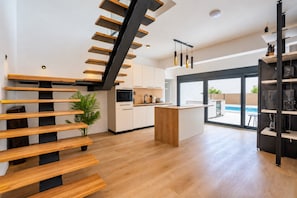 A modern kitchen with a central island, wooden staircase, and a view of a swimming pool.