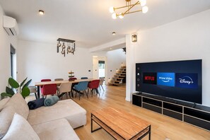 A modern living room with a flat-screen TV, a dining area with chairs, and a staircase leading to another level.