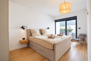 A modern bedroom with a large bed, a bedside table, a chair, and a view of the outdoors through a large window.