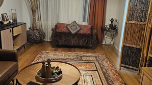Een woonkamer met een ronde salontafel, een bank, een kroonluchter en een groot raam met gordijnen.