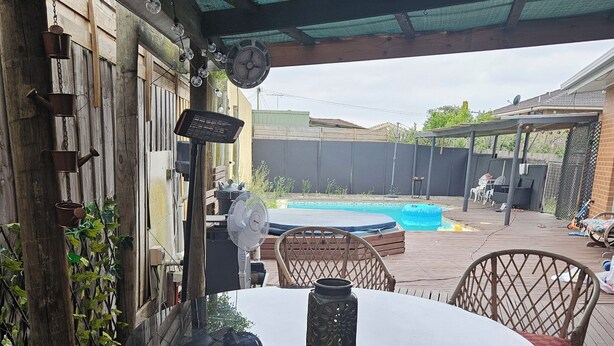 Een overdekt terras buiten met een glazen tafel, rieten stoelen en een zwembad op de achtergrond.
