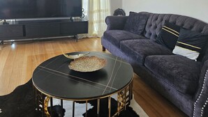Een woonkamer met een donkere bank, een ronde salontafel, een flatscreen televisie en een plafondventilator.