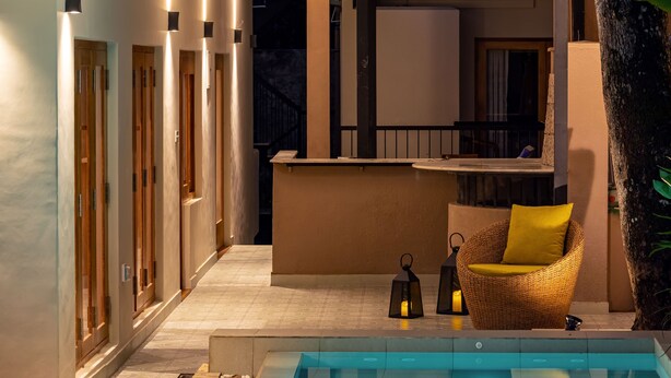 Un'area piscina con bordo curvo, una sedia in vimini con cuscino giallo e un angolo bar con ringhiera.