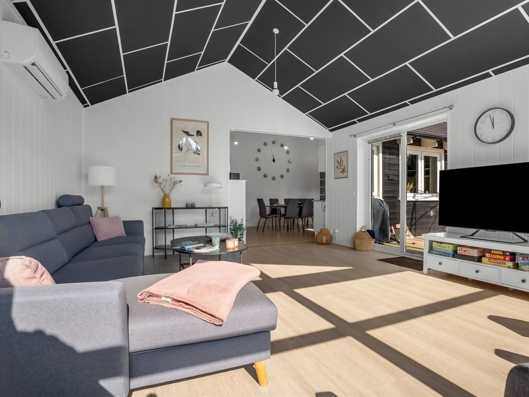 A modern living room with a grey sofa, a coffee table, a flat-screen TV, and a dining area with a clock on the wall.