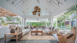 A spacious living area with a high ceiling, large windows, and a view of lush greenery outside.