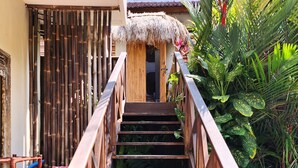 Uma escadaria de madeira leva a uma entrada com telhado de palha, cercada por vegetação exuberante e uma piscina.