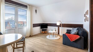 A modern living room with a sofa, a TV, a dining table, and a view of buildings outside. - Apartment 'Inselliebe' with Balcony and Wi-Fi (Pellworm)