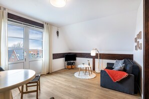 A modern living room with a sofa, a TV, a dining table, and a view of buildings outside. - Apartment 'Inselliebe' with Balcony and Wi-Fi (Pellworm)