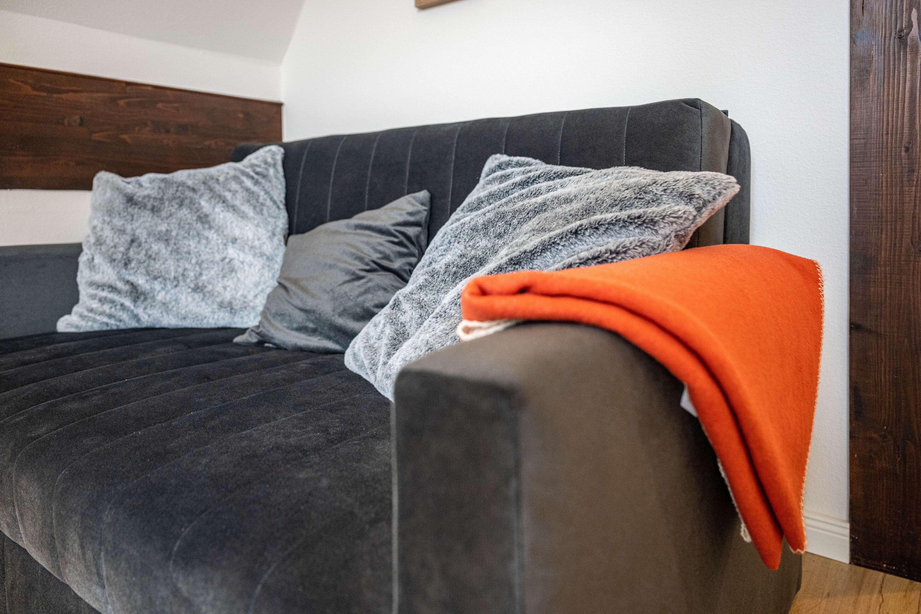 A dark-colored sofa with a grey and orange blanket, two grey pillows, and a wooden headboard.