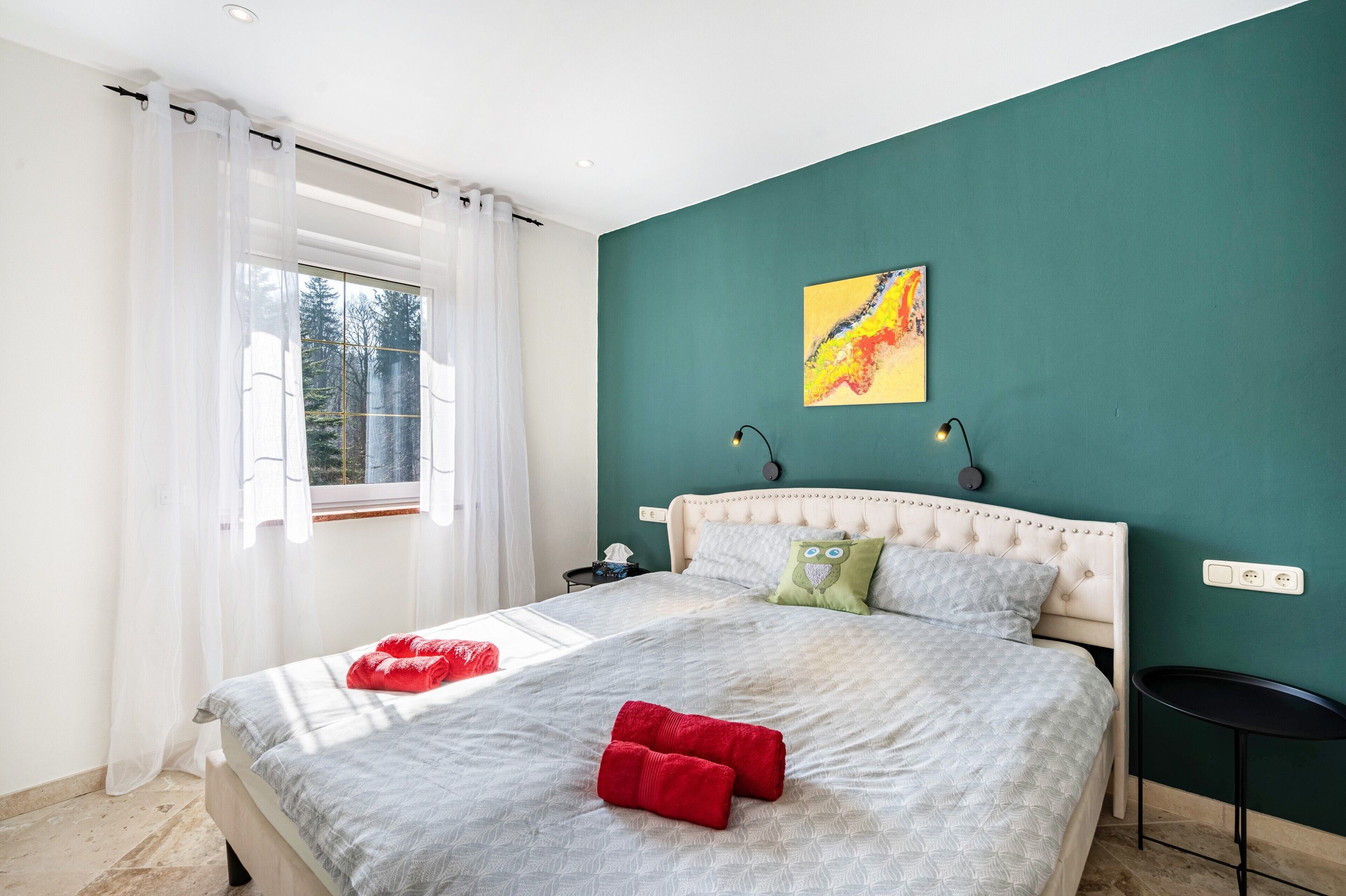 Een net opgemaakt bed met kussens, een nachtkastje, een raam met gordijnen en een muur met een schilderij.
