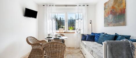 Una sala de estar moderna con un sofá, dos sillas de mimbre, una mesa de comedor, un televisor montado en la pared y un ventanal con vistas.