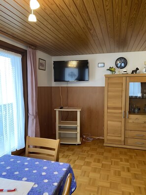 Huoneessa on puinen katto, seinälle asennettu televisio, puinen lipasto laatikoineen, pieni puinen pöytä, jonka päällä on sininen pöytäliina, sekä tuoli.