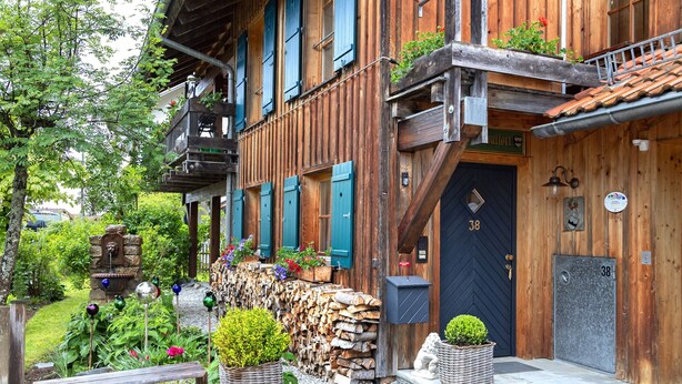 Ein hölzernes Gebäude mit einer schwarzen Tür, auf der '38' steht, einer Holzbank und einem Garten mit Pflanzen und einem Vogelbad.