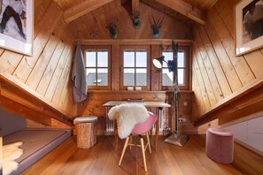 Un intérieur en bois comprenant une table à manger, une chaise recouverte d’un plaid en fourrure, un tabouret et un petit pouf rond.