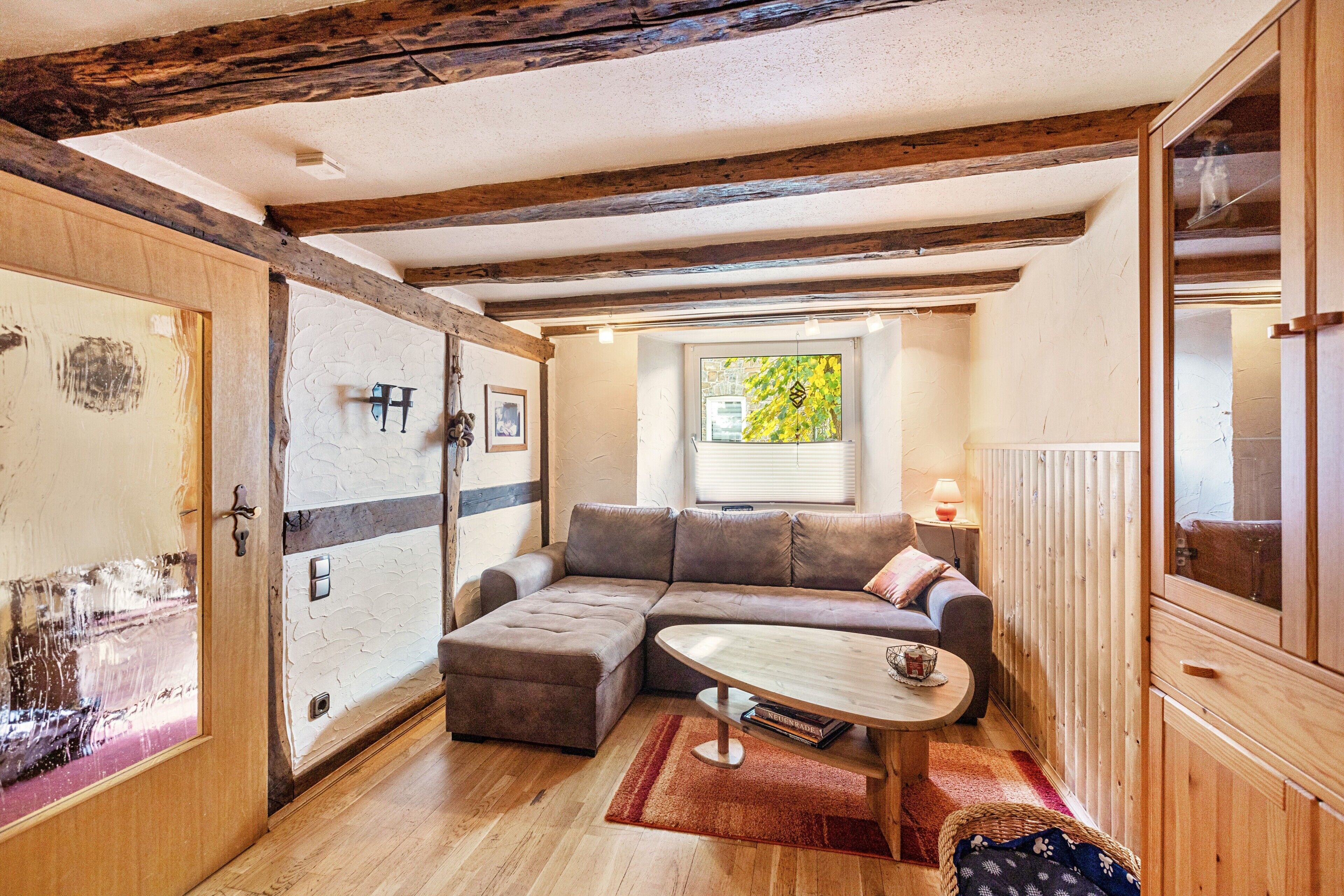 Een knus ingerichte kamer met een houten plafond, een hoekbank, een klein rond tafeltje en een raam met uitzicht op groen.