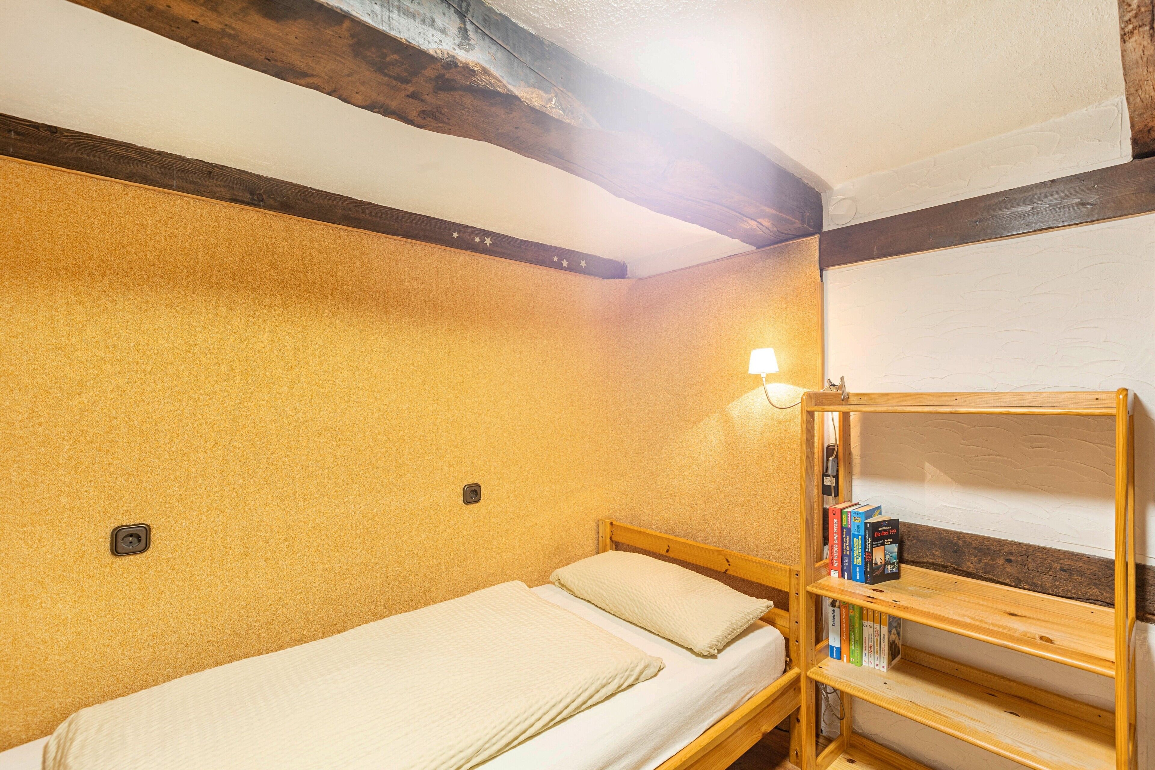 A single bed with a wooden headboard, a shelf with books, and a wall-mounted light fixture.