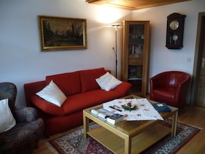 Un salon avec un canapé rouge, un fauteuil à motifs, une table basse avec des livres et une télécommande, une bibliothèque et une horloge murale.